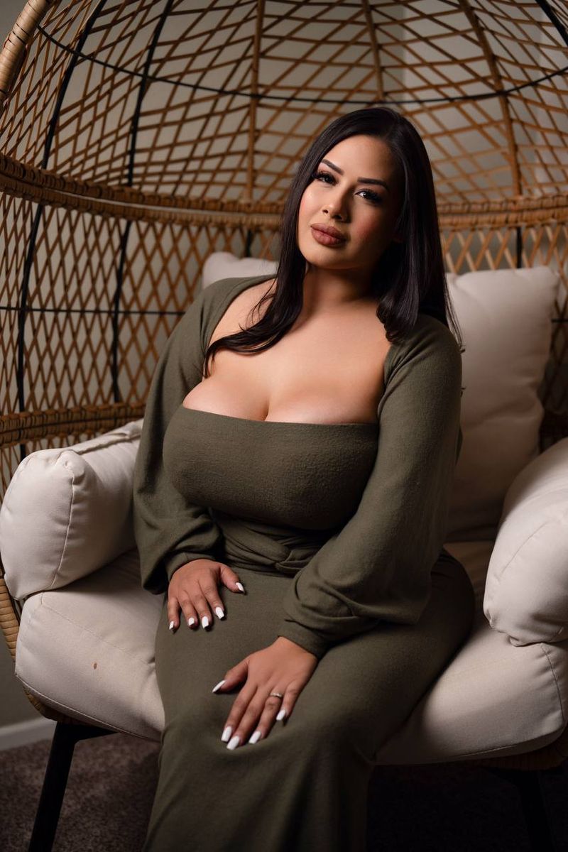 Alivia smiling softly in a rattan egg chair, olive green dress, cream cushions behind her
